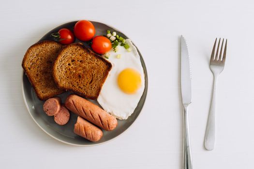 Breakfast plate with knife and fork on the white table, grilled sausage and whole wheat toast with fried egg and cherry tomatoes on plate