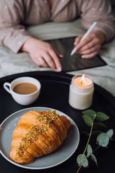 Croissant with cup of cappuccino and scented candle on tray, woman drawing on digital tablet on background