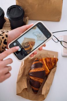 Woman taking photos of her lunch on smartphone, delicious croissants and cups of coffee on workplace, food delivery