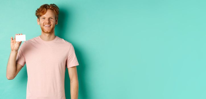 Handsome young man smiling and showing plastic credit card, standing in t-shirt against turquoise background.