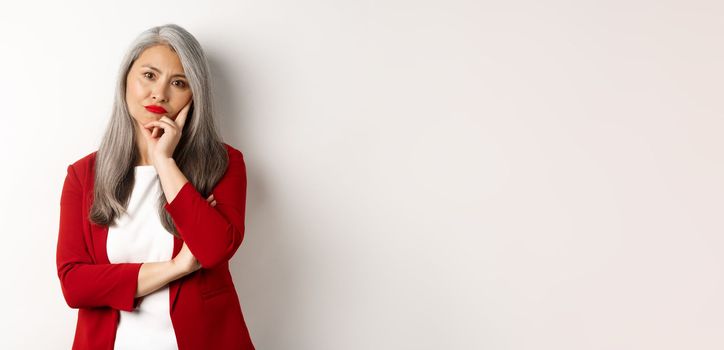 Annoyed and bothered asian businesswoman in red blazer, pouting and looking irritated at camera, standing over white background.