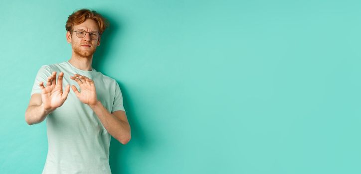 Reluctant redhead man in glasses raising hands in refusal gesture, step backwards, avoiding something with disgusted and unamused face, standing over turquoise background.
