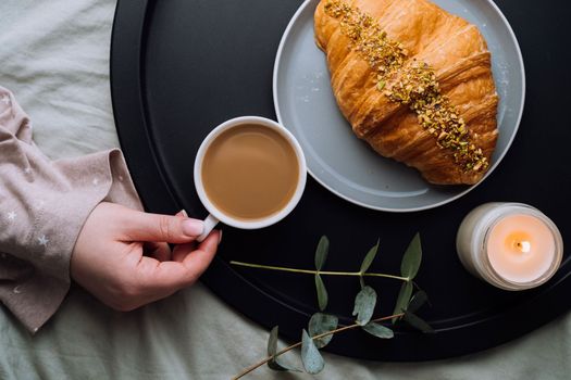 Flat lay of woman's hand holding cup of cappuccino, croissant and scented candle with eucalyptus branch on tray
