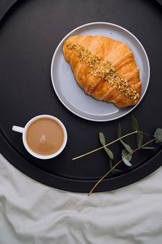 Flat lay of cup of cappuccino, croissant and eucalyptus branch on tray on bed