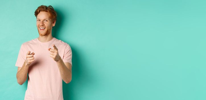 Funny redhead guy congratulating you, pointing fingers at camera and smiling, making choice, standing in t-shirt over mint background.