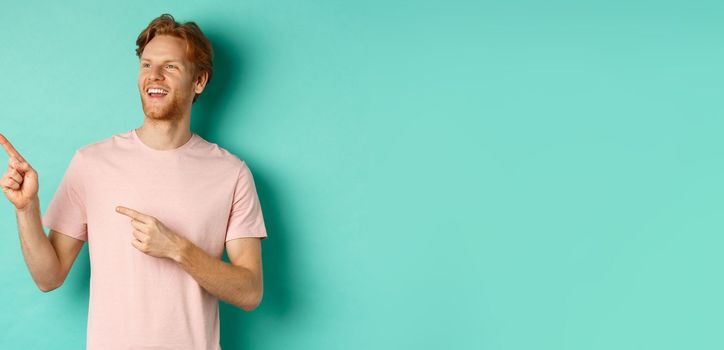 Dreamy handsome guy with red hair, smiling and looking at upper left corner with awe, admire something, standing over mint background. Copy space