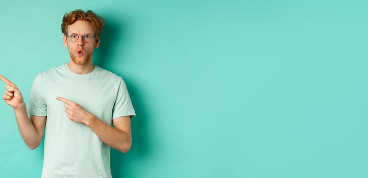 Surprised redhead guy in glasses and t-shirt pointing fingers left, saying wow and showing promo offer, checking out special deal, standing over turquoise background.