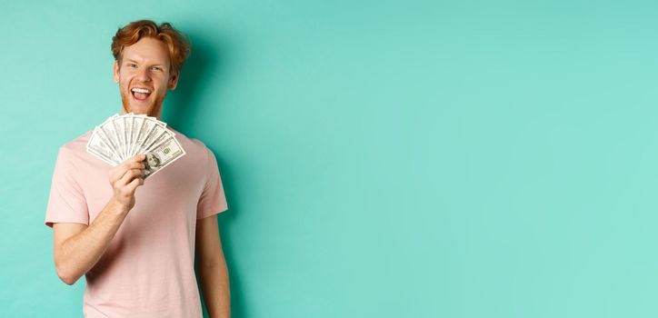 Happy redhead man in t-shirt showing money in dollars and smiling, making smug faces after winning cash, standing over turquoise background.