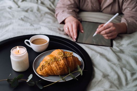 Croissant with cup of cappuccino and scented candle on tray, woman drawing on digital tablet on background