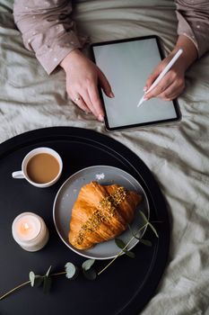 Woman using digital tablet for drawing while lying on the bed and enjoying croissant with cappuccino