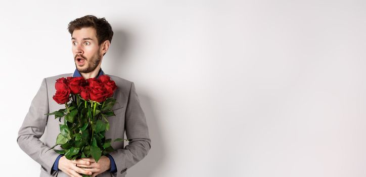 Handsome young man in suit holding red roses, looking left with surprised and startled expression, standing on Valentines day over white background.