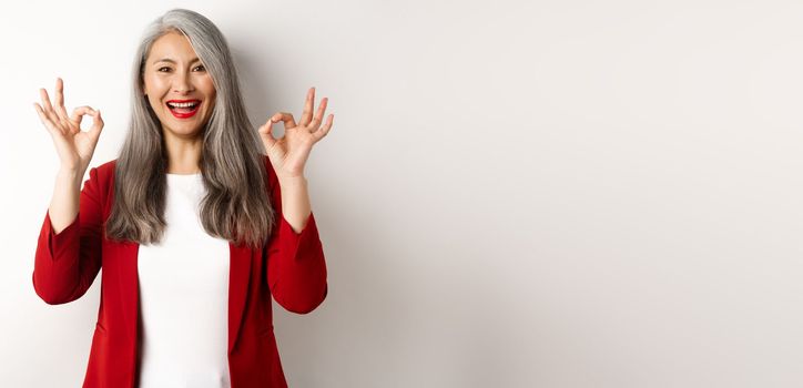 Beautiful senior asian woman in red blazer, smiling and showing okay signs, approve and say yes, standing over white background.