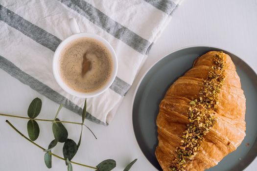 Flat lay of cup of cappuccino with kitchen towel, croissant and eucalyptus branch on table