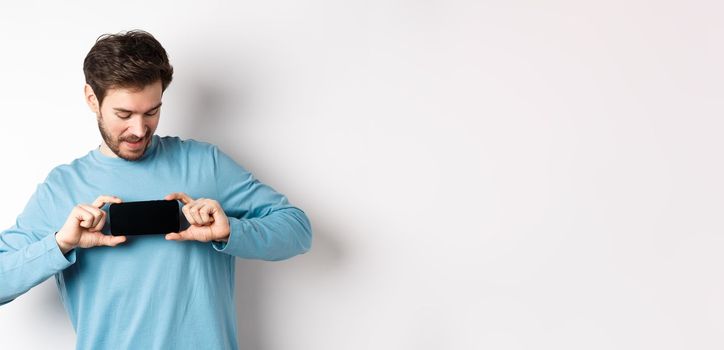 E-commerce and shopping concept. Caucasian man showing blank mobile screen and looking at it, standing curious against white background.