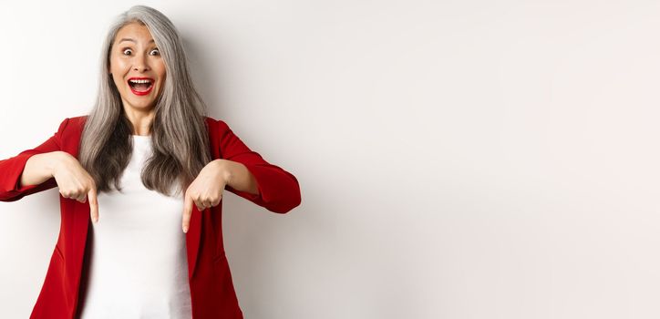 Portrait of happy asian lady in red blazer showing logo, pointing fingers down and smiling cheerful, check this out gesture, white background.