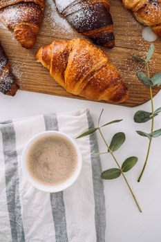 Flat lay of cup of cappuccino with kitchen towel and eucalyptus branch, brown croissants on cutting board