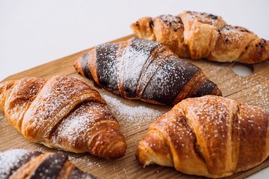 Close up bunch of appetizing brown and chocolate croissants with powdered sugar on wooden board on white table