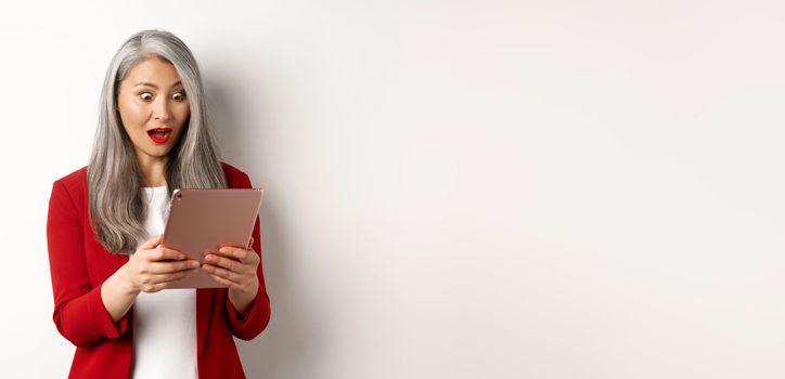 Business. Asian senior woman staring at digital tablet screen with amazed and surprised face, reading amazing news online, standing over white background.