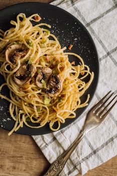 Spaghetti pasta with champignon mushrooms sprinkled with cheese parmesan on a black plate with a towel and a fork on wooden table