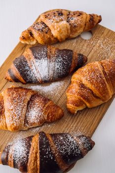 Close up bunch of appetizing brown and chocolate croissants with powdered sugar on wooden board on white table