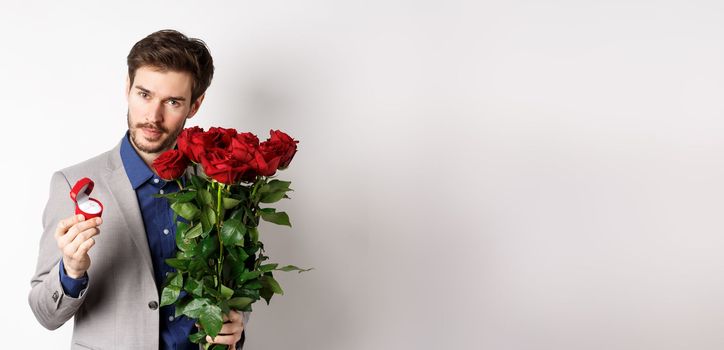 Handsome man in suit, showing engagement ring and looking romantic at camera, standing with red roses over white background. Valentines day and love concept.