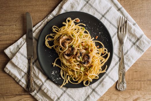 Spaghetti pasta with champignon mushrooms sprinkled with cheese parmesan on a black plate with a towel and a fork on wooden table