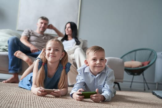 Children sister and brother playing using phone and digital tablet on floor while young parents relaxing at home on sofa.