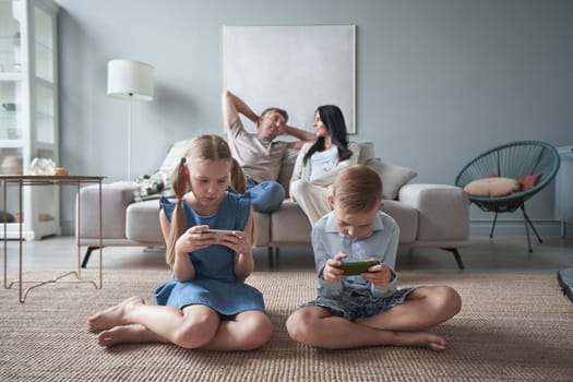 Children sister and brother playing using phone and digital tablet on floor while young parents relaxing at home on sofa.