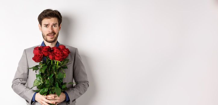 Worried boyfriend in suit, holding flowers roses and looking doubtful at camera, standing with bouquet on valentines day against white background.
