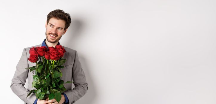 Charming smiling man going on romantic date, wearing suit, holding bouquet of roses and looking left, standing on valentines day with gift on white background.