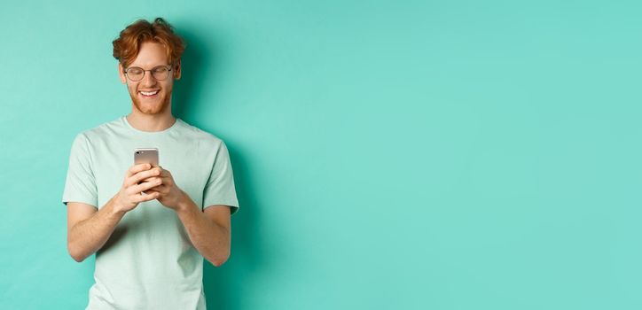 Handsome young man in glasses with red messy hair reading message on mobile phone, smiling and looking at screen, standing over mint background.