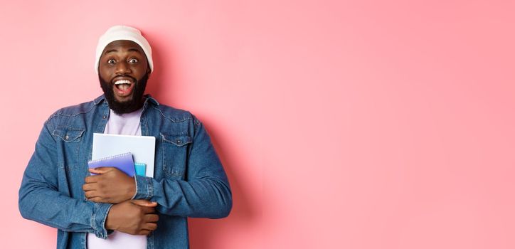 Image of adult african-american man holding notebooks and smiling, studying at courses, standing over pink background.