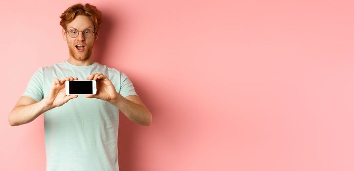 Amazed redhead man gasping and staring with awe phone, showing blank smartphone screen horizontally, standing over pink background.