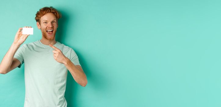 Shopping concept. Handsome redhead man in t-shirt showing plastic credit card and smiling, standing over turquoise background.