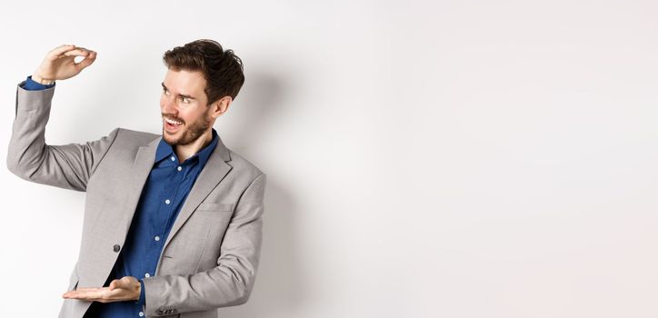 Smiling happy businessman in suit showing large size object, shaping big thing with hands and look pleased, standing on white background.