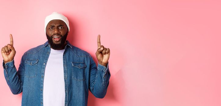 Disappointed and upset Black man frowning, pointing fingers up and staring at camera bothered, complaining at something, standing over pink background.