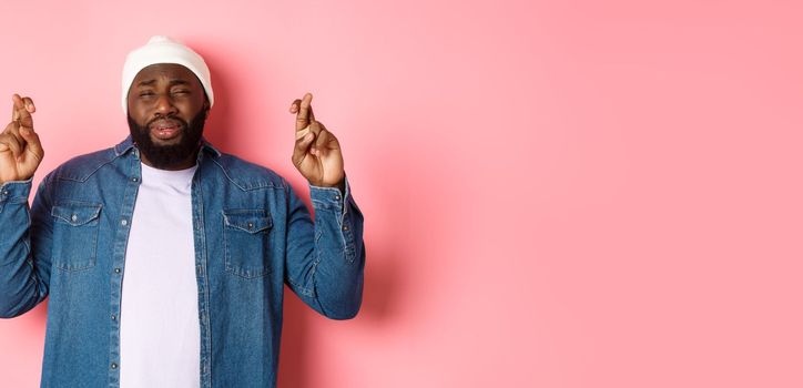 Image of young african-american man whining, pleading god, cross fingers and crying, standing over pink background.