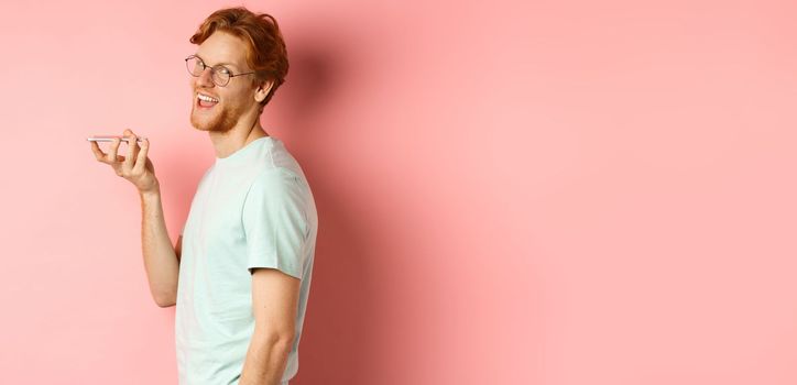 Handsome young man standing in profile and talking on speakerpone, record voice message, turn head at camera and smiling pleased, standing over pink background.