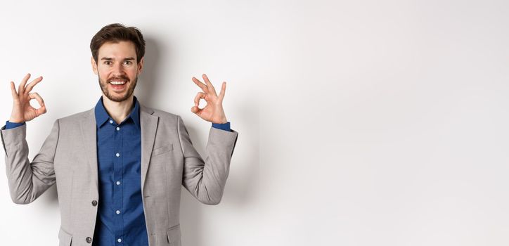 Good choice. Smiling businessman in suit showing okay signs and look happy, recommending good deal, like and approve your choice, standing satisfied on white background.
