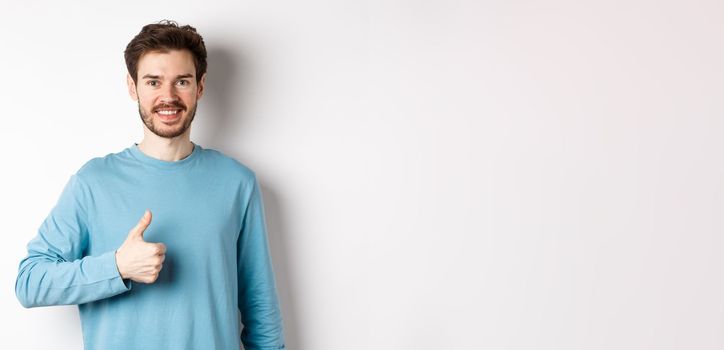 Smiling male model in casual shirt showing thumb up, approve and recommend product, praise excellent choice, standing on white background.