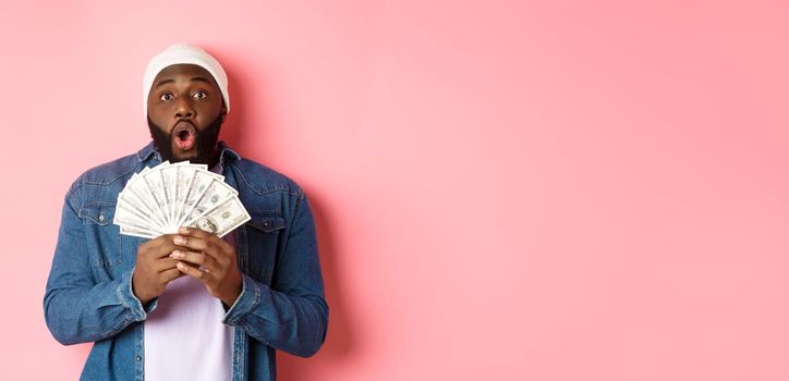 Amazed african-american man receive cash prize, showing money and staring at camera in awe, standing over pink background.