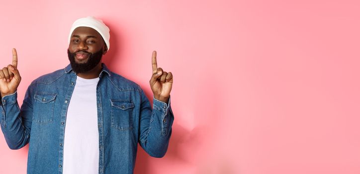 Pleased handsome Black man nodding in approval and checking out good promo, pointing fingers up at offer, showing best deal, pink background.