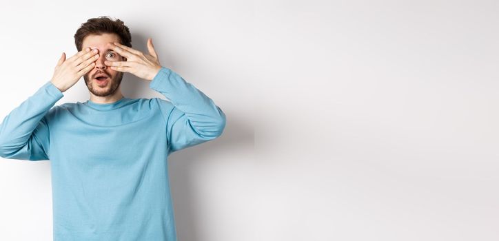 Amazed smiling man covering eyes with hands but peeking through fingers at something awesome, standing over white background.