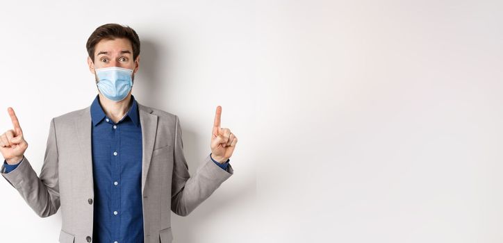 Covid-19, pandemic and business concept. Excited businessman in sterile medical mask and suit pointing fingers up, white background.