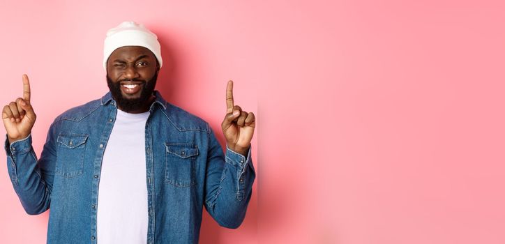 Cheeky african american man smiling and winking, showing deal on top, pointing fingers up, standing over pink background.