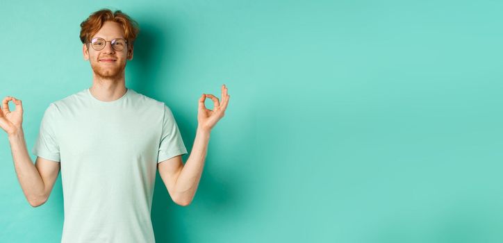 Calm and relaxed redhead man in glasses smiling, holding hands spread sideways with om mudra gesture, meditating peaceful, standing over mint background.