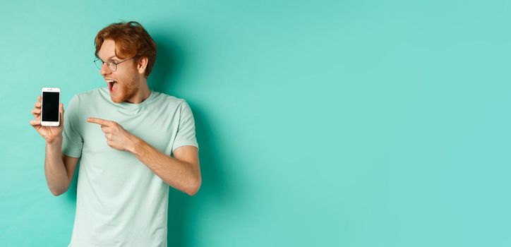 Check this out. Handsome redhead guy in glasses pointing finger at blank smartphone screen, showing online promotion, standing amazed over turquoise background.