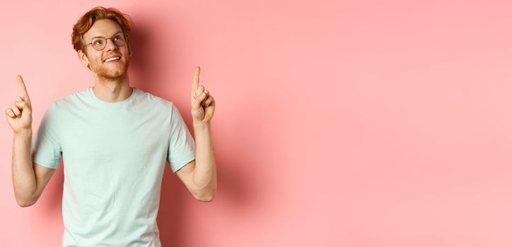Dreamy young man with red hair and beard pointing fingers and looking cheerful up, admire something, standing over pink background.
