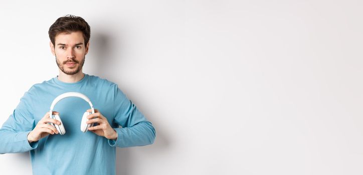 Young bearded guy in blue sweatshirt put on wireless headphones, listening music, standing on white background.