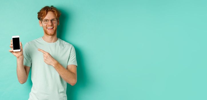 Attractive young man with red beard and hair pointing finger at blank smartphone screen, showing online promotion or app, smiling at camera, turquoise background.